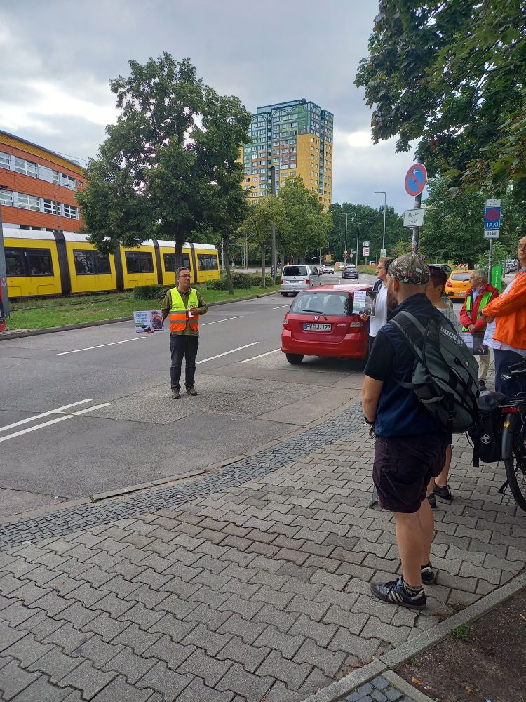 Mahnwache mit verschiedenen Personen die für eine Schweigeminute auf der Fahrbahn sitzen. Im Hintergrund fährt eine Tram.