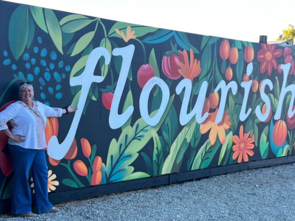 A mural with fruits, vegetables and flowers in the background and the word “flourish” in the foreground. I am standing next to the approximately 8 ft tall mural with my hand on my hip. 