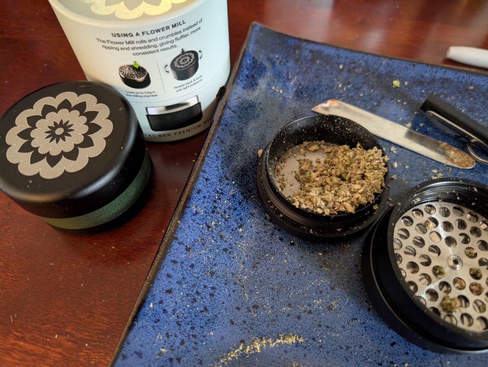 A picture of a closed Flower Mill with a green anodized ring (where we keep our daytime herbs). The box with the instructions on how to use the grinder. An open Flower Mill with herbs (not oregano) ground coarsely. We see a scoop made by an Edmonton blacksmith and a Pax Packing tool on a blue plate/ceramic tray.