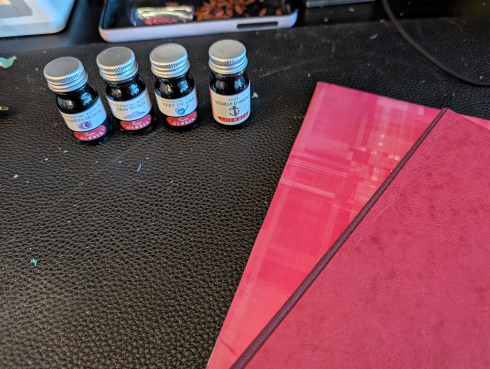 Four ink bottles with 10ml of ink are lined up. Colours are Poussière de lune (purple), Gris nuage (grey), vert de gris (green), and ancre de rouille (pink). 

Two soft cover notebooks in pink and red can also be seen.

The desk where the picture is taken is a little messy, with a lot of paper pills and cables all over