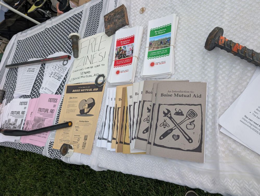 A zine table with boise mutual aid zines and various other zines. Heavy paper weights can be seen holding the lighter things down, this is foreshadowing