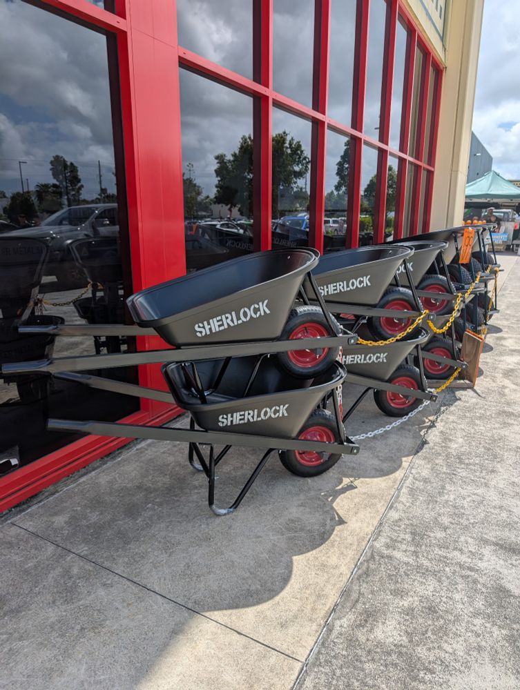 A row of stacked black wheelbarrows in front of a hardware store window with the brand name Sherlock painted in white on each.