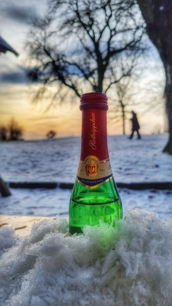 Sektfläschchen im Schnee (Frost) im Hintergrund verschwommen warmes Licht mit Baum und einer gehender Person. 