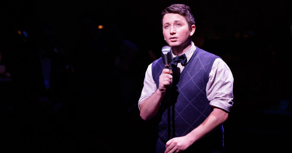 A photo of B.J. Gibson, 1940s Radio Hour, Hillbarn Theatre, December 2021. 

A man standing a microphone, wearing a button up shirt with faint stripes, a blue sweater, and a bow tie.