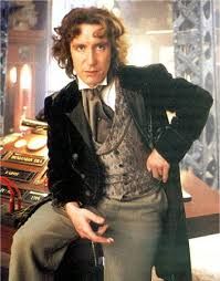 Photo of actor Paul McGann as the 8th Doctor in Doctor Who. He wears a dandyish 19th century type of outfit, including a paisley silk or satin waistcoat, a velvet frock coat and a cravat with a tie pin.
