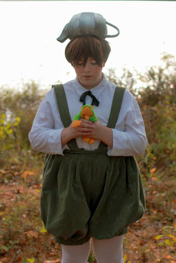 Greg from Over the Garden Wall, from the knees up in front of fall foliage and a washed out lake. Looking down at the frog plushie they hold in their hands. 