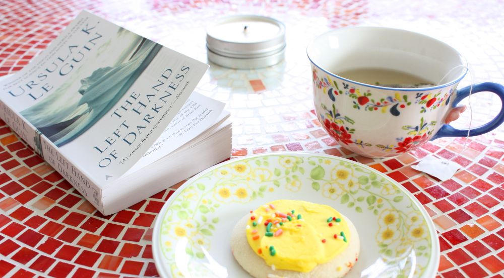 The Left Hand of Darkness by Ursula K. Le Guin on a table with a yellow frosted cookie and a floral tea cup.