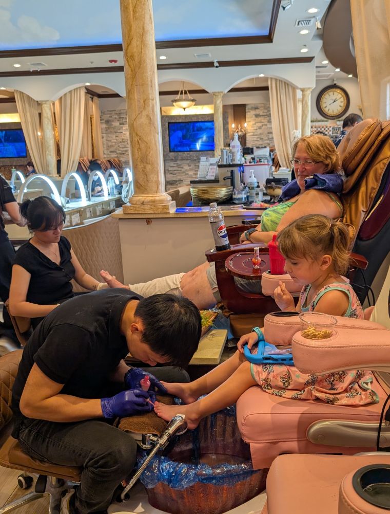 Interior of a nail salon. A lady is sitting next to a four year old girl while both are being attended to.  The girl has animal crackers in her mouth and a somewhat skeptical look.