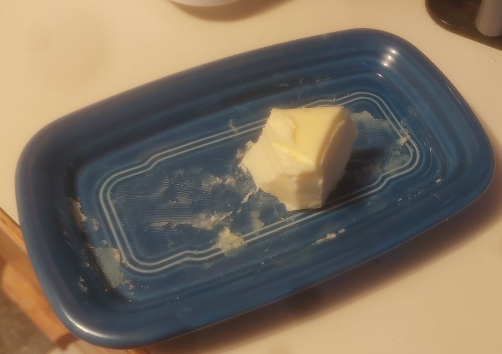 A butter dish containing the sad remains of a stick of butter. About two tablespoons are left of the stick but people have been taking bits off from at least five distinct angles, none of which are at a right angle to the length of the original stick.