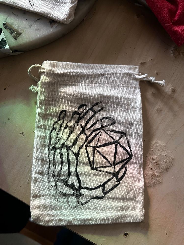 A photo of a canvas cinch bag with a block print of a skeleton hand holding a d20