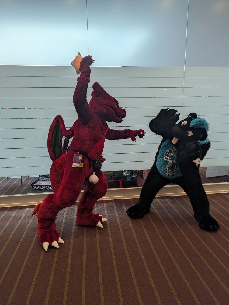 Charkol (he/him), a charizard fursuit, is posing with Kiba (she/they), a blue and black skunk. Charkol is swinging the poptart overhead and is preparing to smack Kiba with it while she holds up her paws cowering