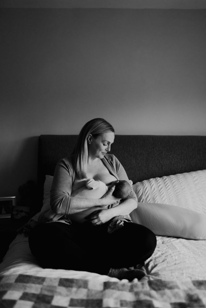 Black and white image of a white woman breastfeeding while sitting on a bed. 
