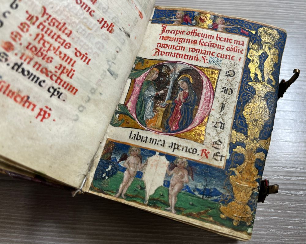 The first page of Matins, with an historiated initial showing the Annunciation and a full-page border with gold architectural ornaments and putti in front of a mountainous landscape.
