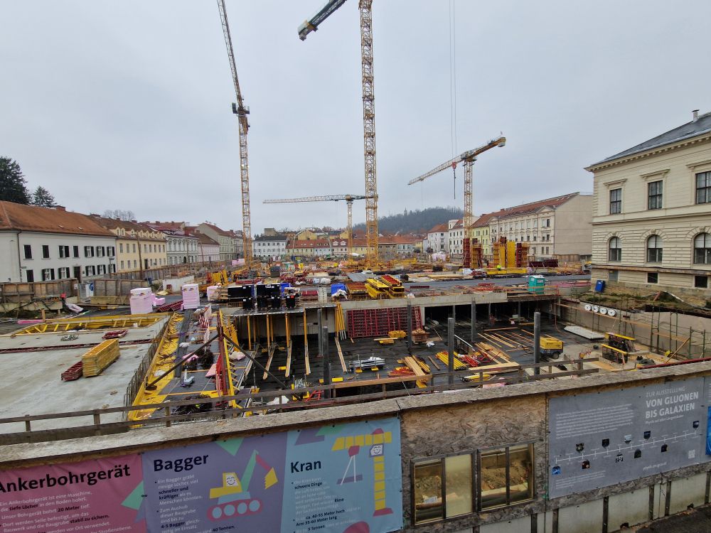 A large construction site with multiple cranes. The constructed building is almost level with the surrounding roads almost everywhere. Its overcast.