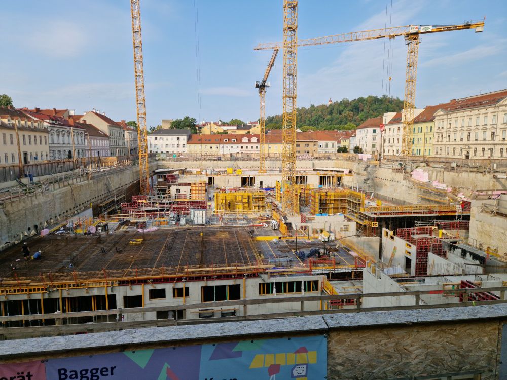 A very large construction site with multiple cranes. The lowest base floor is completely made from concrete, and first rooms are visible, including already some ceilings. It is sunny.