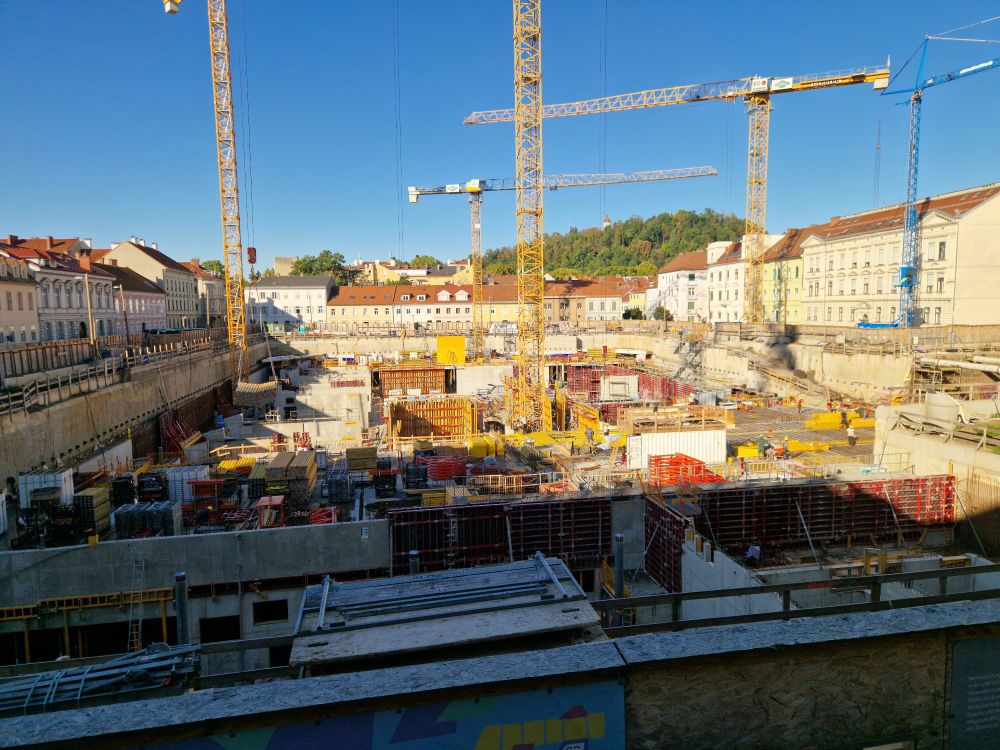 A large construction site below street level. Multiple structures visible, and multiple cranes operating. It is sunny.
