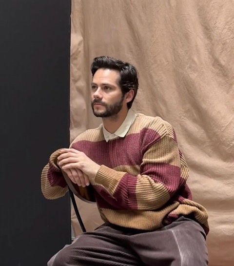 Dylan O’Brien on the photoshoot set of Entertainment Weekly wearing a striped sweater and brown corduroy pants. He is posed sideways on a chair with his hands resting atop of the backrest. 