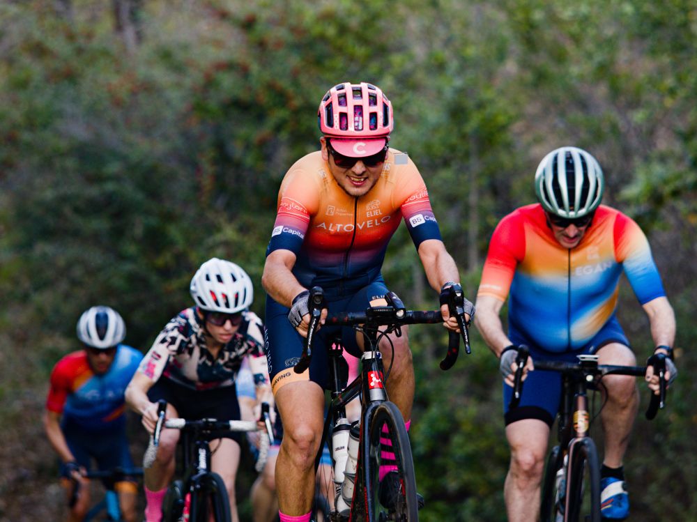 Jonathan is riding his bike with a pained face up a hill with other cyclists in the background. 