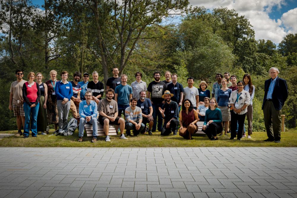A group of about twenty people is standing and sitting on a grassy area in front of trees and a blue sky. Some are wearing name tags.