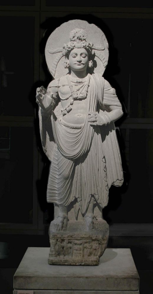 Statue représentant un bodhisattva sous forme humaine datée entre le Ier et IIIème siècle, réalisée au Gandhara et conservée au musée Guimet à Paris. Le bodhisattva se tient debout, légèrement souriant, la main gauche posée sur la hanche et la droite légèrement levée devant lui, comme en signe de bienvenue. Il porte une fine moustache et sur sa tête une sorte de couronne épaisse au sommet de laquelle se trouve une forme ronde évoquant un chignon. Il est habillé d'un large pantalon de toile tandis qu’une étoffe tombant de son épaule gauche laisse découverte la majeure partie de son torse. Divers bijoux et ornements pendant se son cou et de ses épaules. Sa tête est auréolée d'un cercle plein sur lequel figurent un motif en demi cercle ainsi que deux motifs évoquant des cornes placés de chaque côté de sa tête.
Source : https://commons.wikimedia.org/wiki/File:Bodhisattva_Gandhara_Guimet_181171.jpg?uselang=fr