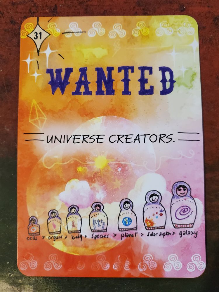 Cosmic Journey oracle card 31 Wanted Universe Creators. Image is a colorful sky with a moon, sun, & clouds. At the top left corner is a diamond with 31 inside and black streaks spanning out from the shape. A yellow upside-dwon kite is under three white four point stars. Along the top and bottom of the card is a border of triskelions in white gradients. In the middled of the card in blue is the word wanted in a western style font. Below that in a hand-lettered font in blue is universe creators with two horizontal lines on each side of the phrase. Below that toward the bottom of the card is a set of Russian nesting dolls.From left to right, small to large, the dolls have a word with corrosponding image on each doll, starting with cells>organs>body>species>planet>solar system>galaxy. The colors in the background are shite, yellow, pink, orange, and a splast of green.