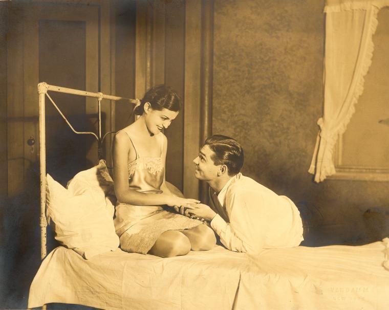 An actress in a nightgown sits on a bed, holding hands w/ a man lying on the bed.