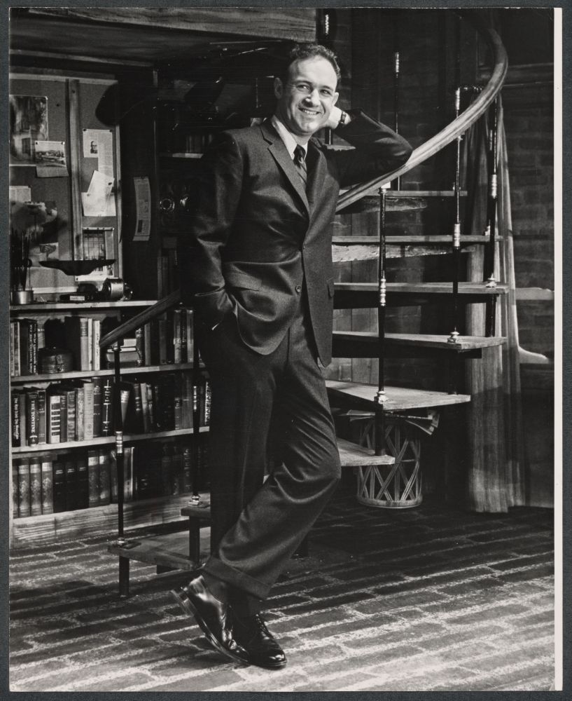 Gene Hackman leans against a staircase, with bookcase in the background. 