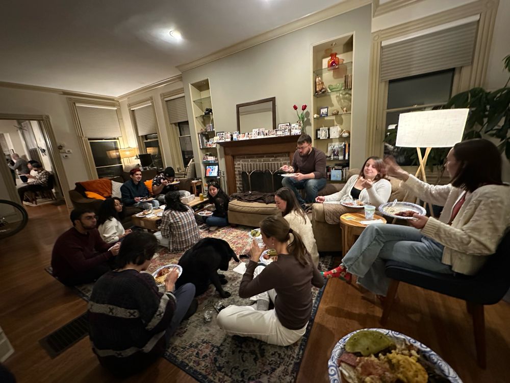 Group of people sitting on the floor or couches in a living room eating food with a black dog in the middle begging for food