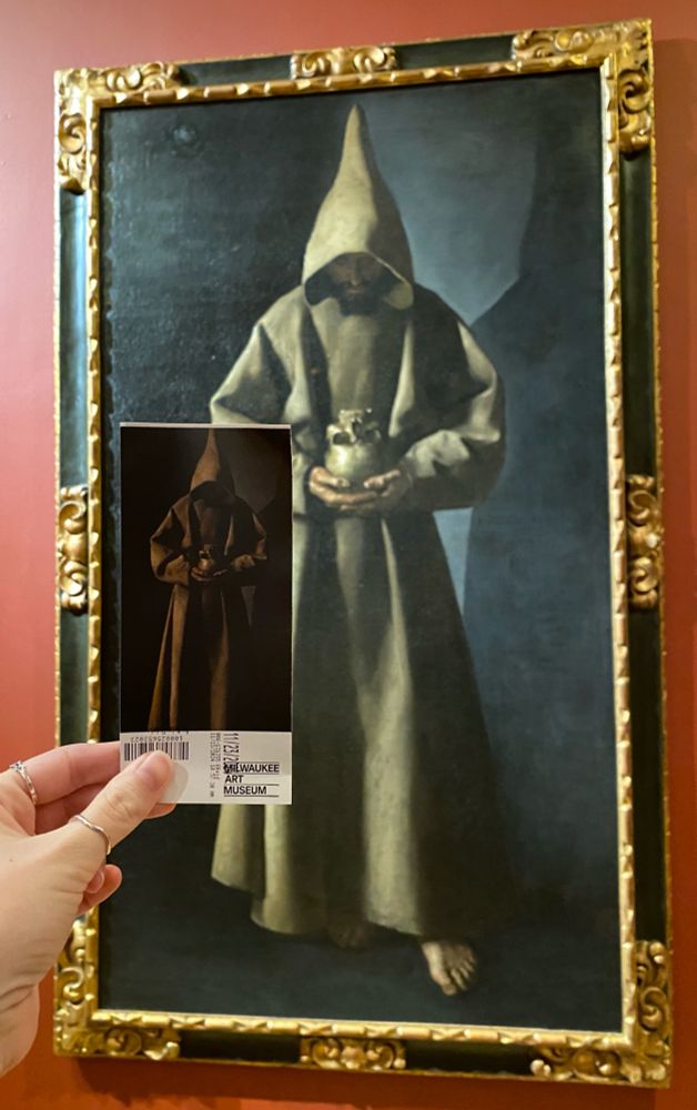 A hand holds a ticket to the Milwaukee Art Museum printed with the seventeenth-century painting Saint Francis of Assisi in His Tomb by Spanish artist Francisco de Zurbarán in front of the actual painting of Saint Francis of Assisi in His Tomb. 
