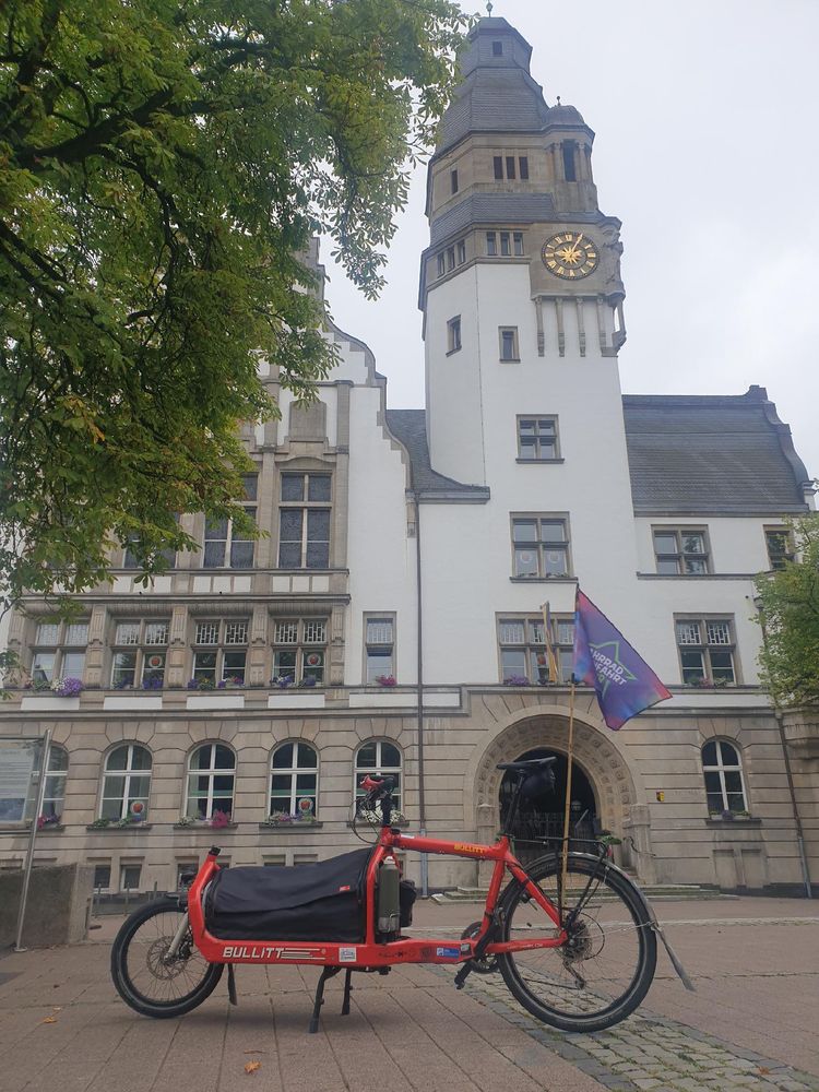 Auf dem Foto ist ein rotes Lastenfahrrad der Marke „Bullitt“ zu sehen, das auf einem Platz steht. Am Rad ist eine bunte Fahne mit der Aufschrift „FahrradSternfahrt.Ruhr“ befestigt. Im Hintergrund befindet sich ein großes, helles Gebäude mit Turmuhr und hohen Fenstern, geschmückt mit Blumen. Vorne ragt ein grüner Baum ins Bild.