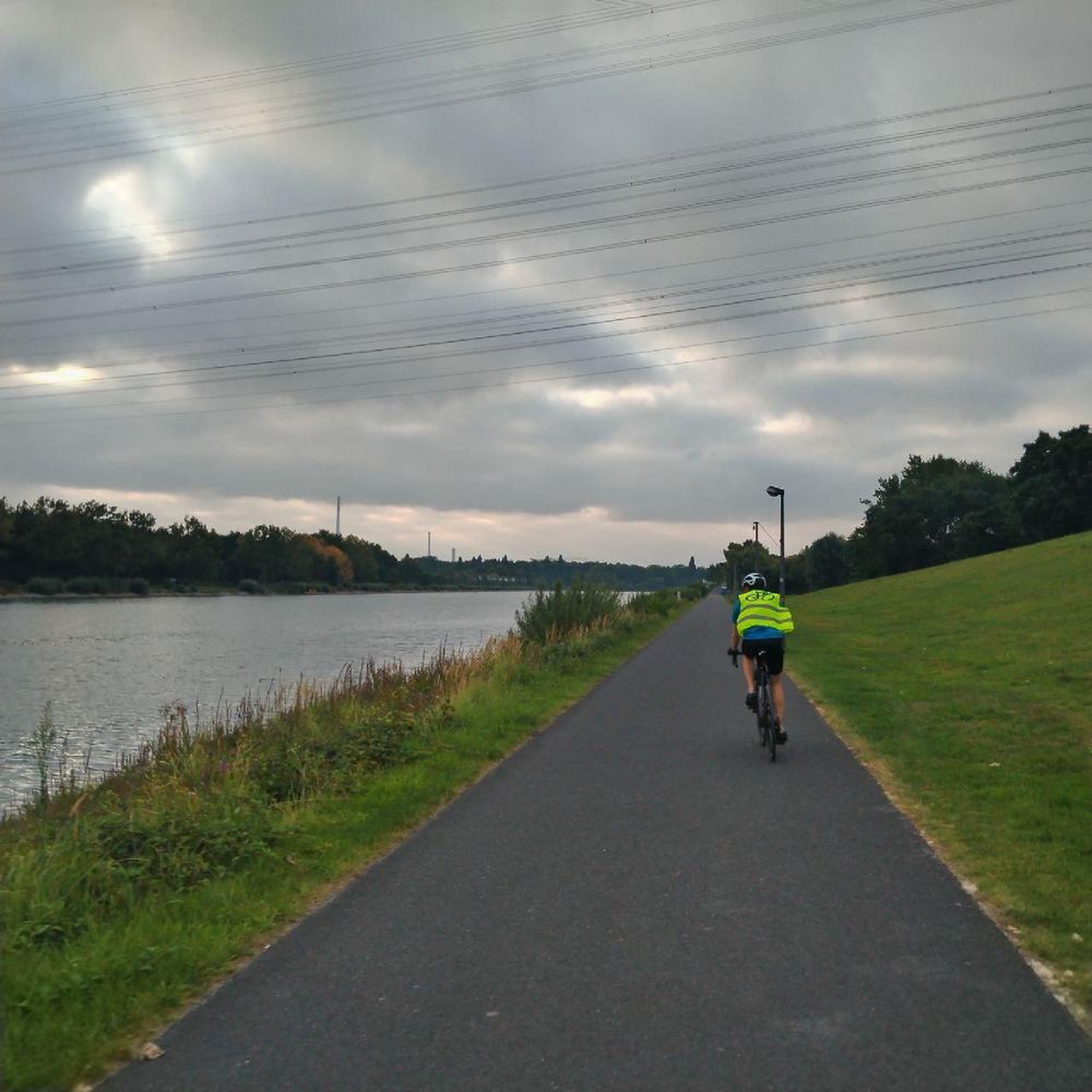 Foto eines Fahrradweges, auf der eine Person mit Warnweste von der Kamera weg radelt. Links ein Fluss, rechts Wiese. Wolkiges Morgenwetter. Am Wegesrand stehen Straßenlaternen.