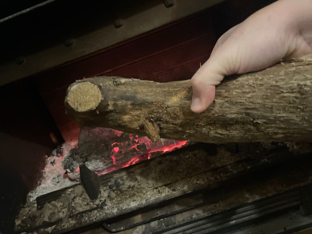 Photo d’une cheminée pleine de braises, dans laquelle on voit ma main mettre une bûche.