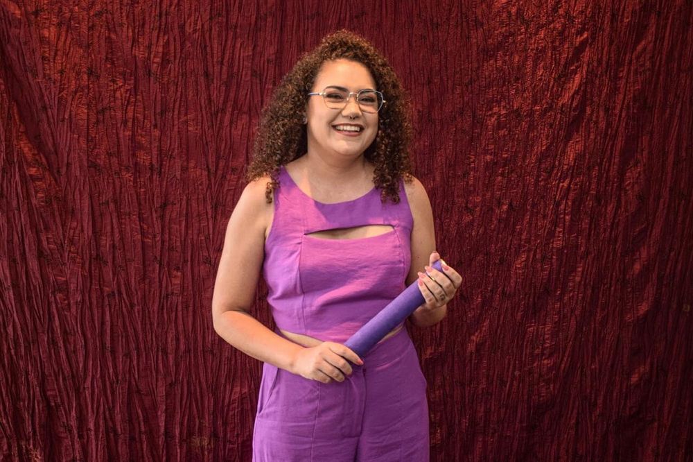 Pessoa de pele branca vestindo blusa e calça lilás, segurando um canudo de formatura. Atrás uma parede vermelha.