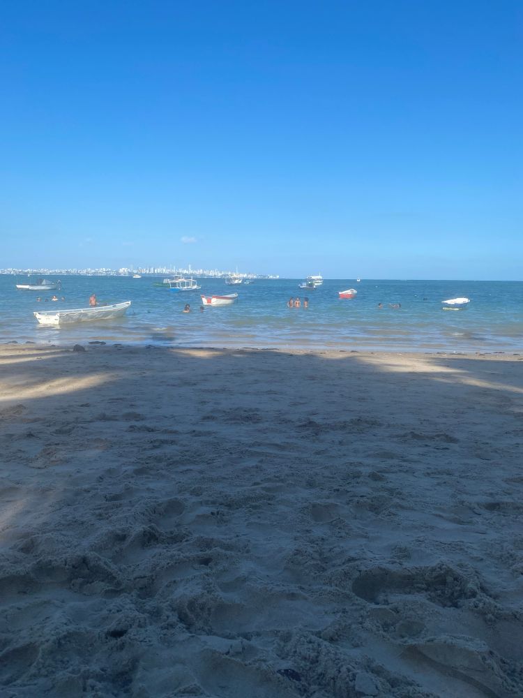 Praia com areia com bastante sombra e com alguns barcos e pessoas na água e céu azul 