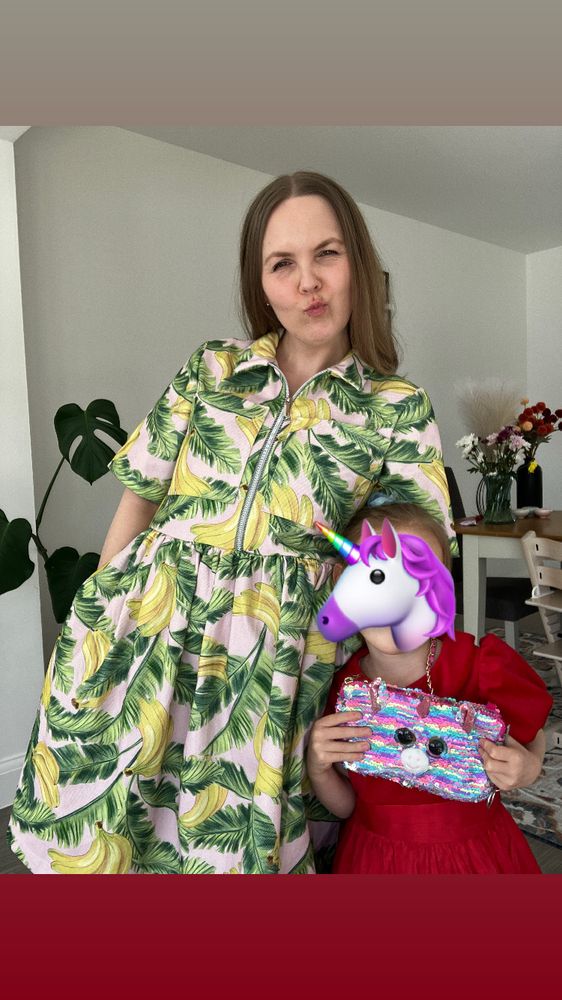 A white woman is facing the camera and wearing a pink denim dress with green leaves and yellow bananas. The dress has short sleeves, breast pockets, a front zip, gathered skirt and is knee length. 