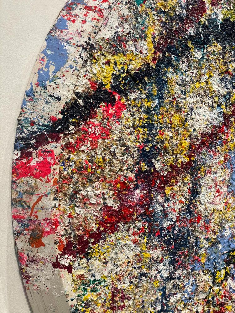 The edge of Sam Gilliam's painting "Lucky" (2021) at the Hirshhorn Museum. A thickly painted and splattered abstract tondo painting, with fluorescent hues covering a white background, with an angled, beveled white frame edge onto which the composition spills.