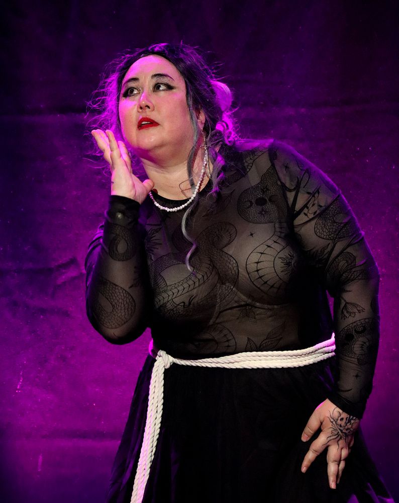 The performer poorlyformed backlit in purple. Their costume consists of a sheer back top with a design reminiscent of Ed's tattoos, a pearl necklace, a black/grey ombre wig (tied back) and a rope around their waist. their right hand (as seen) is on their hip, while their left is up near their face. They gaze off into the upper left corner, looking at something we cannot see.