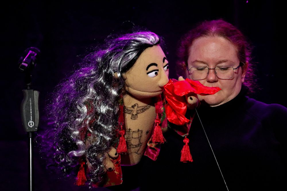 a photo of Rattie & muppet Ed from their performance at The New Uni-Con. The muppet Ed is on the left, holding the piece of red fabric to his face, and wearing the depression robe outfit. Rattie wears theatre black. Both look sombre.