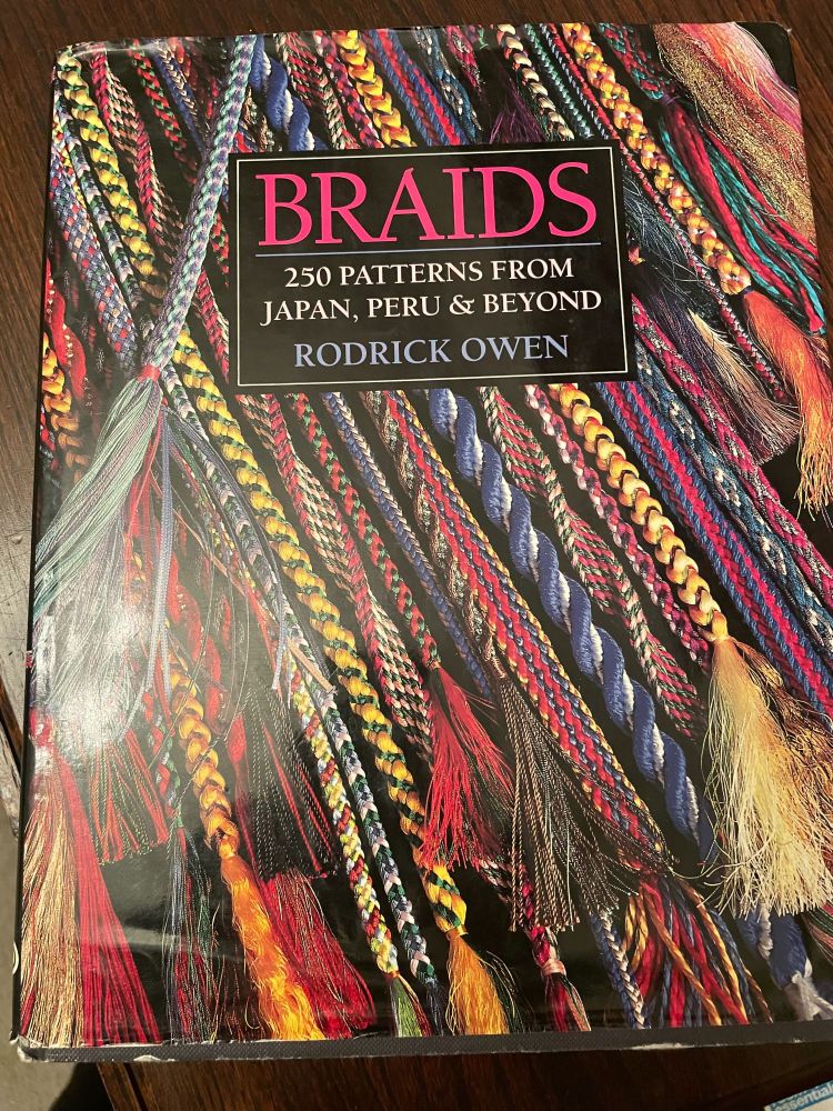The cover of nonfiction book Braids 250 Patterns from Japan Peru & Beyond by Roderick Owen is covered with many bright multi-colored braids. 