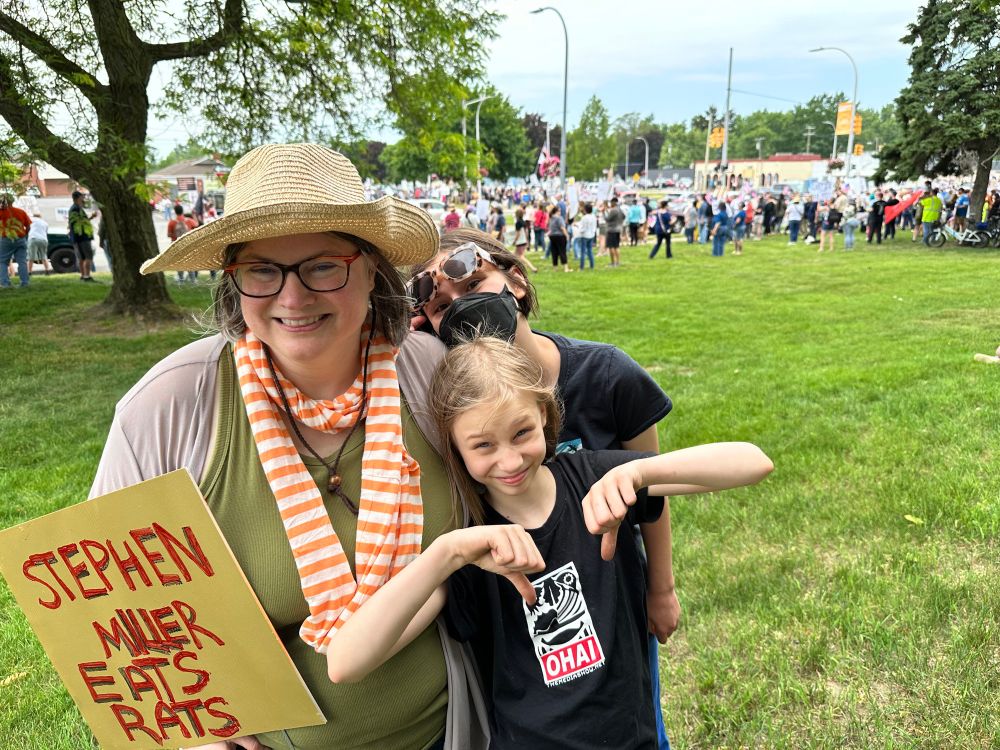 A smiling white lady and her two children pose in front of a crowd of hundreds of protesters. Her sign says “Stephen Miller Eats Rats”. One child give two thumbs down. 