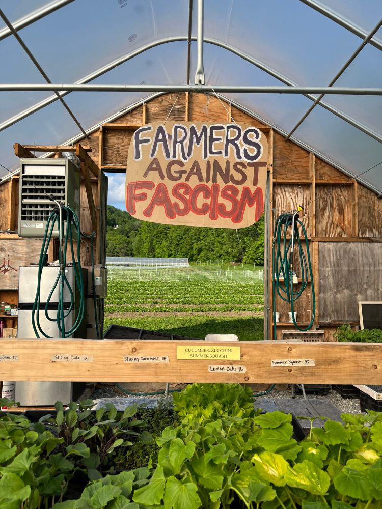Greenhouse with seedlings; farm fields can be seen through the open back door; sign from the ceiling reads: ‘ Farmers against fascism’