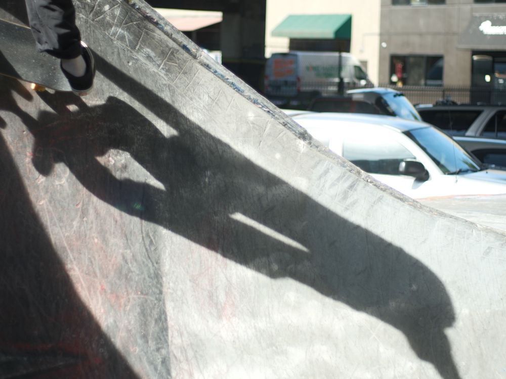 the shadow of a skateboarder stretched and wrapped along the contour of a bowl. cars and light industrial in the background. 
