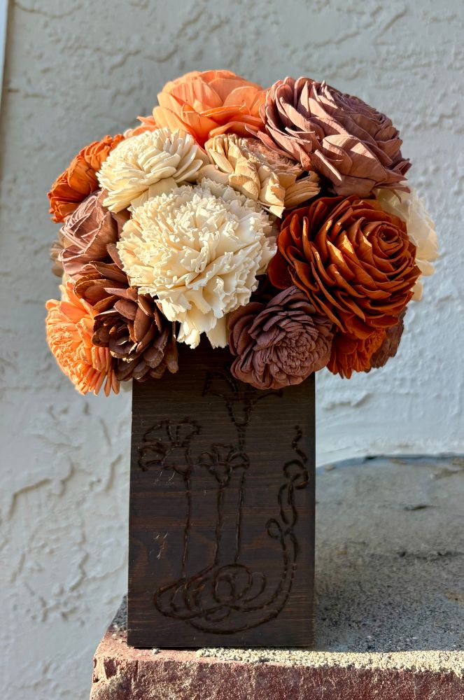 a dark brown, square, wooden vase filled with wooden flowers in shades of orange, brown, and cream. the vase has a art nouveau flowers wood burned into it.