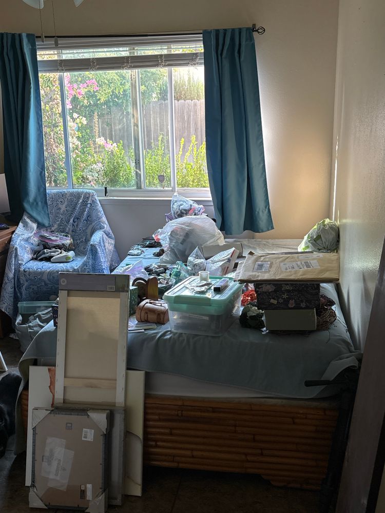 a bedroom featuring a bed covered in miscellaneous heaps of art supplies. there is a stack of canvases on the floor leaning on the bed. a chair next to the bed is also full of supplies 