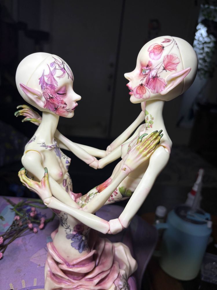 A ball-jointed doll that is two bodies/ribcages connected to one another. The figure on the right is sculpted from the hips up, with below the hips disappearing into a large flower. The figure on the left is sculpted from the hips up, but emerging from the abdomen of the first. 

Both dolls are covered in what look like flower tattoos, the one on the right with more purple the one on the left more pink. The doll on the right has closed eyes and purplish makeup, the doll on the left has open eyes and pink makeup. They both have pointed ears, and long, bony fingers with sharp nails. Both dolls' fingers are tinged green.

The doll is sitting on a rather messy work surface.