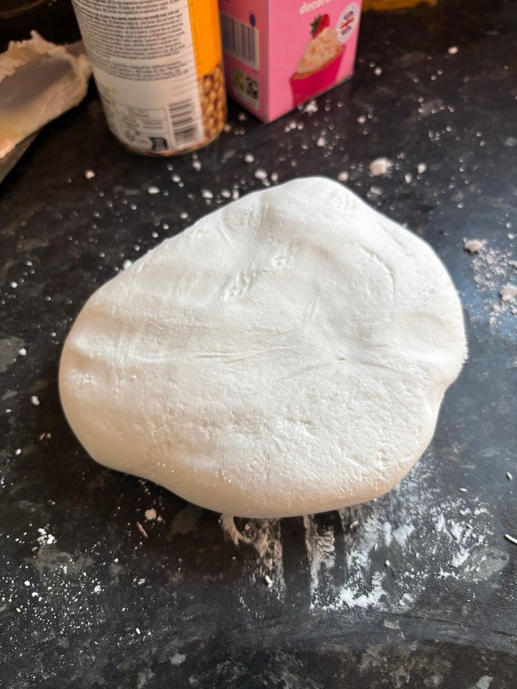 A piece of rolled royal icing shaped into a flat, rounded form sits on a dark countertop dusted with icing sugar. In the background, there are kitchen items