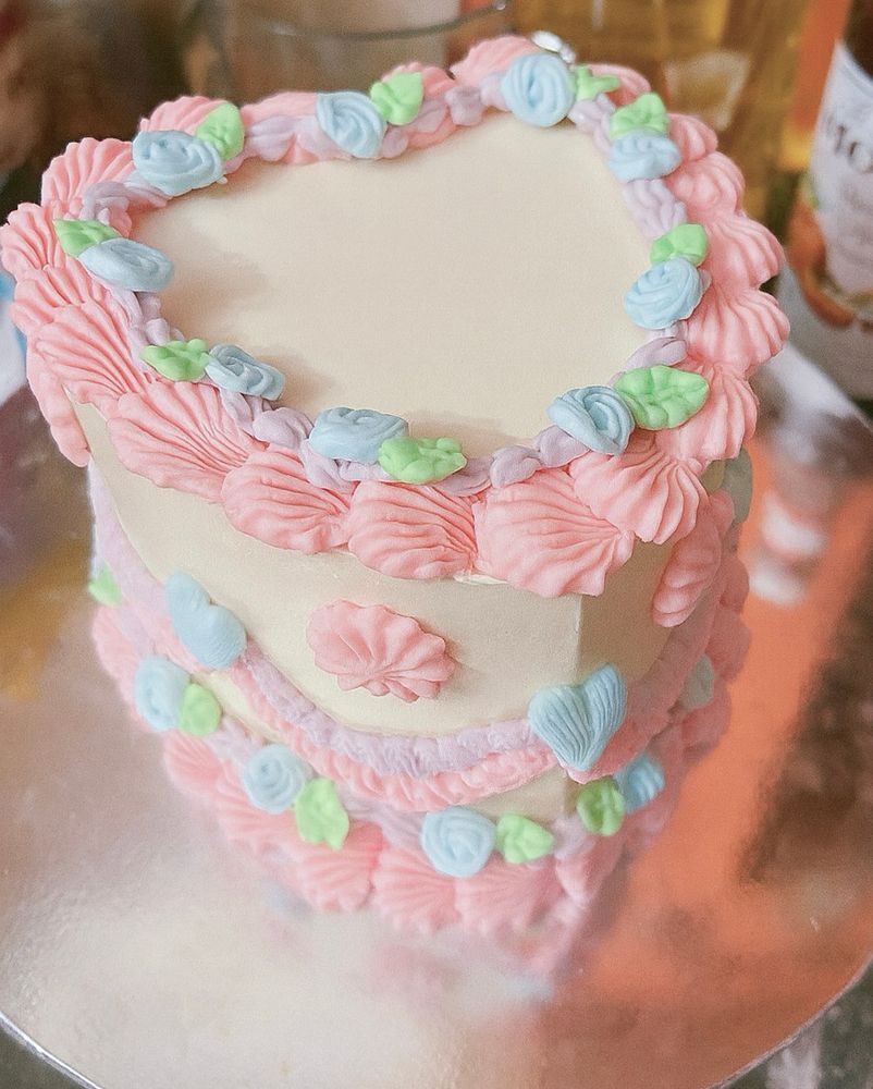 A heart-shaped cake decorated in the Lambeth style. The cake is covered in smooth white icing with piped pastel decorations. Pink shell borders and swirls frame the top and sides, with accents of blue rosettes, green leaves, and lilac details. The cake sits on a reflective silver cake board.