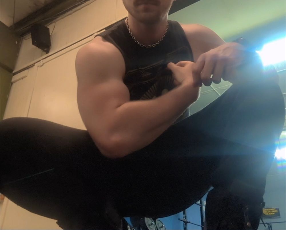 A picture of a caucasian man wearing all black sweatpants and a sleeveless shirt kneeling with the upper half of his face out of frame to center his muscular arm, which is flexed and positioned to create a triangle of space between his chest, forearm, and bicep. 
