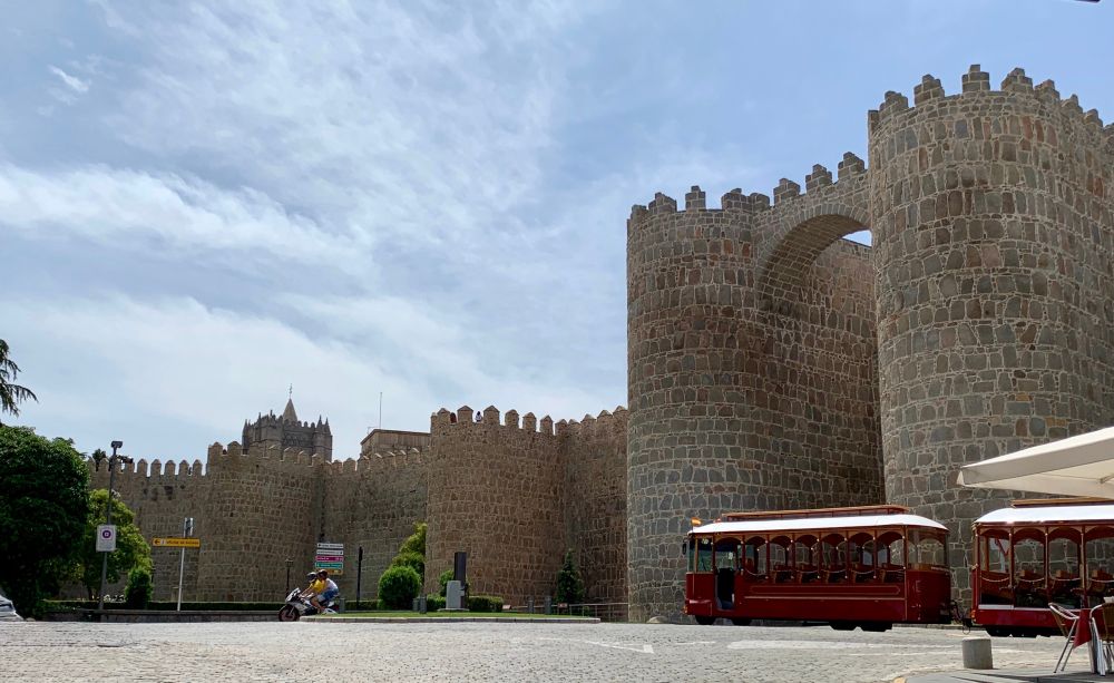 Vista de una puerta y parte de la muralla. A la derecha un tren turístico. Al fondo una pareja circula en motocicleta. Cielo azul y nubes blancas.