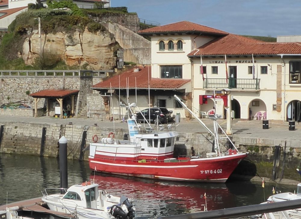 En un puerto pesquero un barco rojo y blanco. Edificios al fondo. Cielo azul.